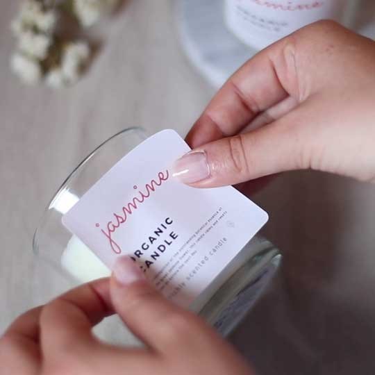 Mãos aplicando um etiqueta com vela orgânica escrita nele em um frasco de vidro transparente